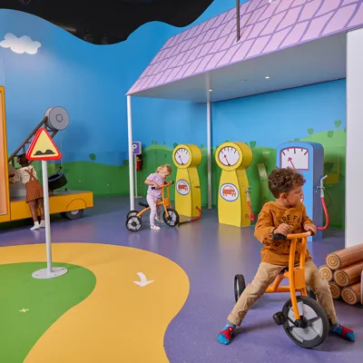 At PEPPA PIG World of Play in Chicago, young children play in Grandad's Garage, a colorful interactive area featuring a pretend gas station and a Peppa Pig-themed bus in a fun indoor setting.